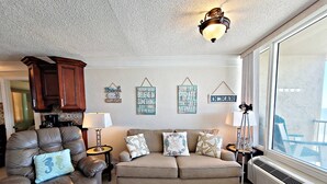 Una sala de estar con un sillón de cuero marrón, un sofá color beige y una mesa de centro. Hay decoraciones en las paredes, una lámpara y una ventana con vistas.