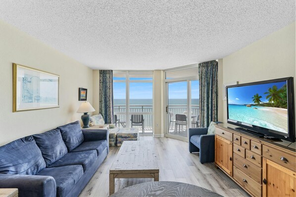 Uma sala de estar com sofá, mesa de centro, televisão e varanda com vista para o mar.