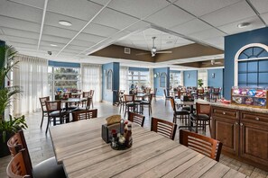 Uma ampla sala de jantar com várias mesas e cadeiras, um balcão de cozinha com diversos utensílios, e janelas grandes que permitem a entrada de luz natural.