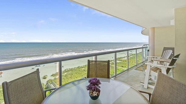 Un balcone con tavolo in vetro, sedie e un vaso di fiori. Si vede l'oceano e una spiaggia.