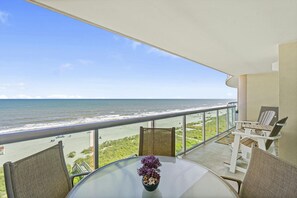 A balcony with a glass table, chairs, and a vase of flowers. The view includes the ocean and a beach.