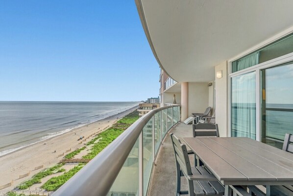 Un balcón con vistas a la playa y al océano, una mesa con sillas y una barandilla de cristal.