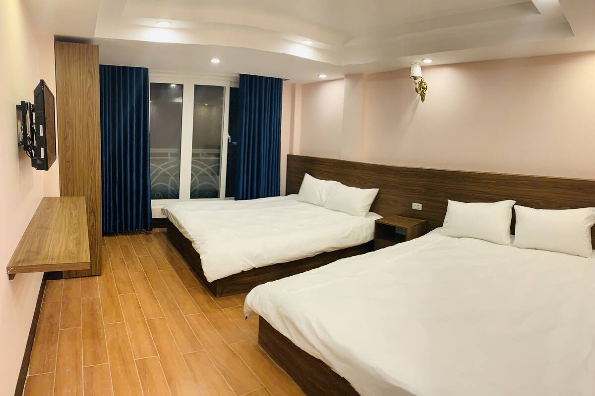 Quarto de hotel com duas camas, cabeceira de madeira, televisão e varanda com cortinas azuis.
