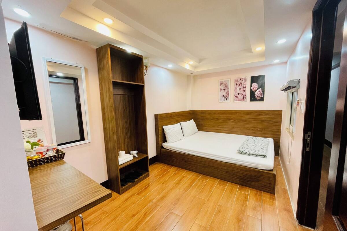 Quarto de hotel compacto com cama de madeira, uma pequena mesa de escritório, guarda-roupa e espelho.