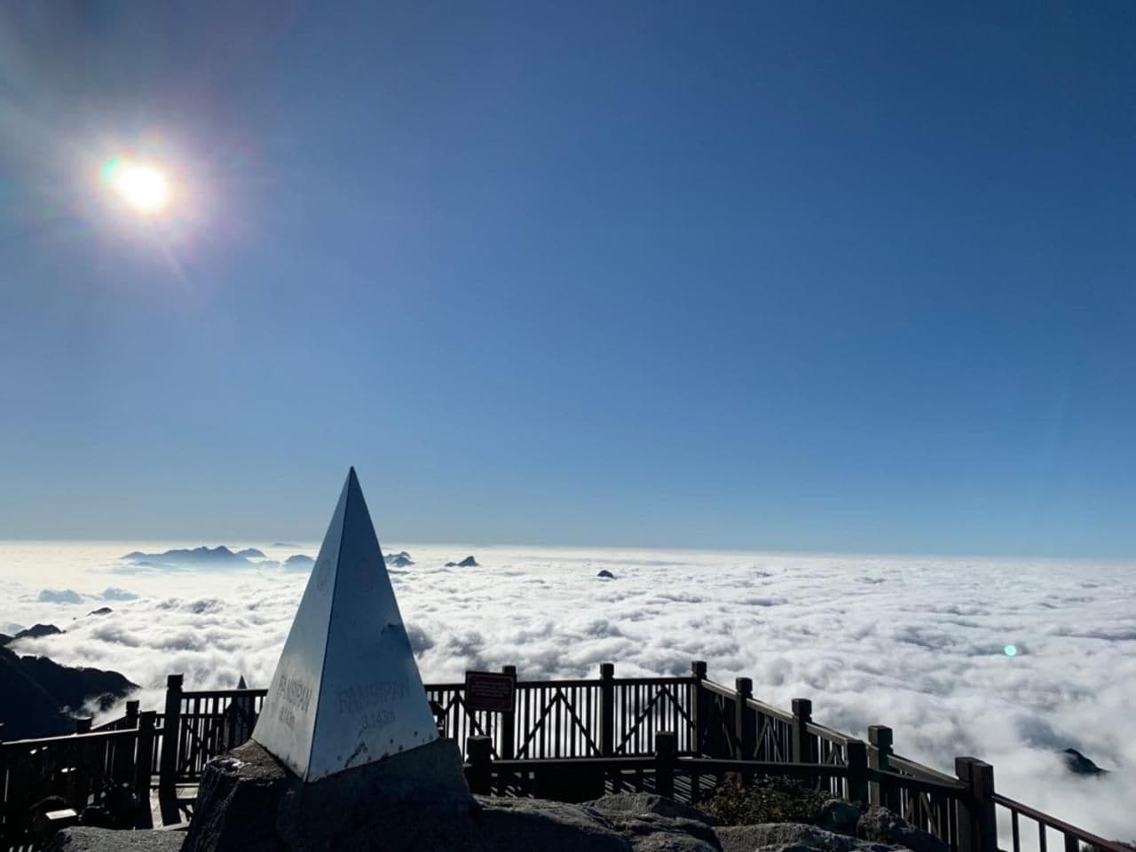 Cielo despejado con el sol brillando, la cima de una montaña y una valla de madera.