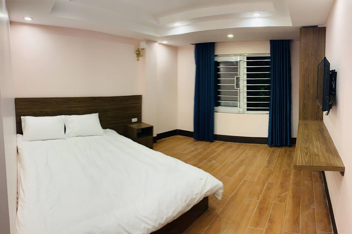Quarto de hotel com uma cama, mesa de cabeceira, televisão, janela com cortinas e piso de madeira.
