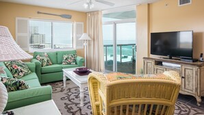 Habitación de hotel con sofá verde, una silla de mimbre, televisor y balcón con vista al océano.