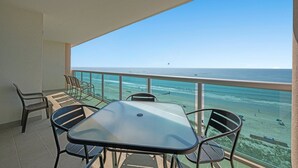Un balcón con mesa y sillas de vidrio, con vista a una playa de aguas azules y una isla distante.