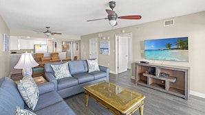 A living room with a sofa, coffee table, and TV. There are ceiling fans, a kitchen in the background, and a beach scene on the TV.