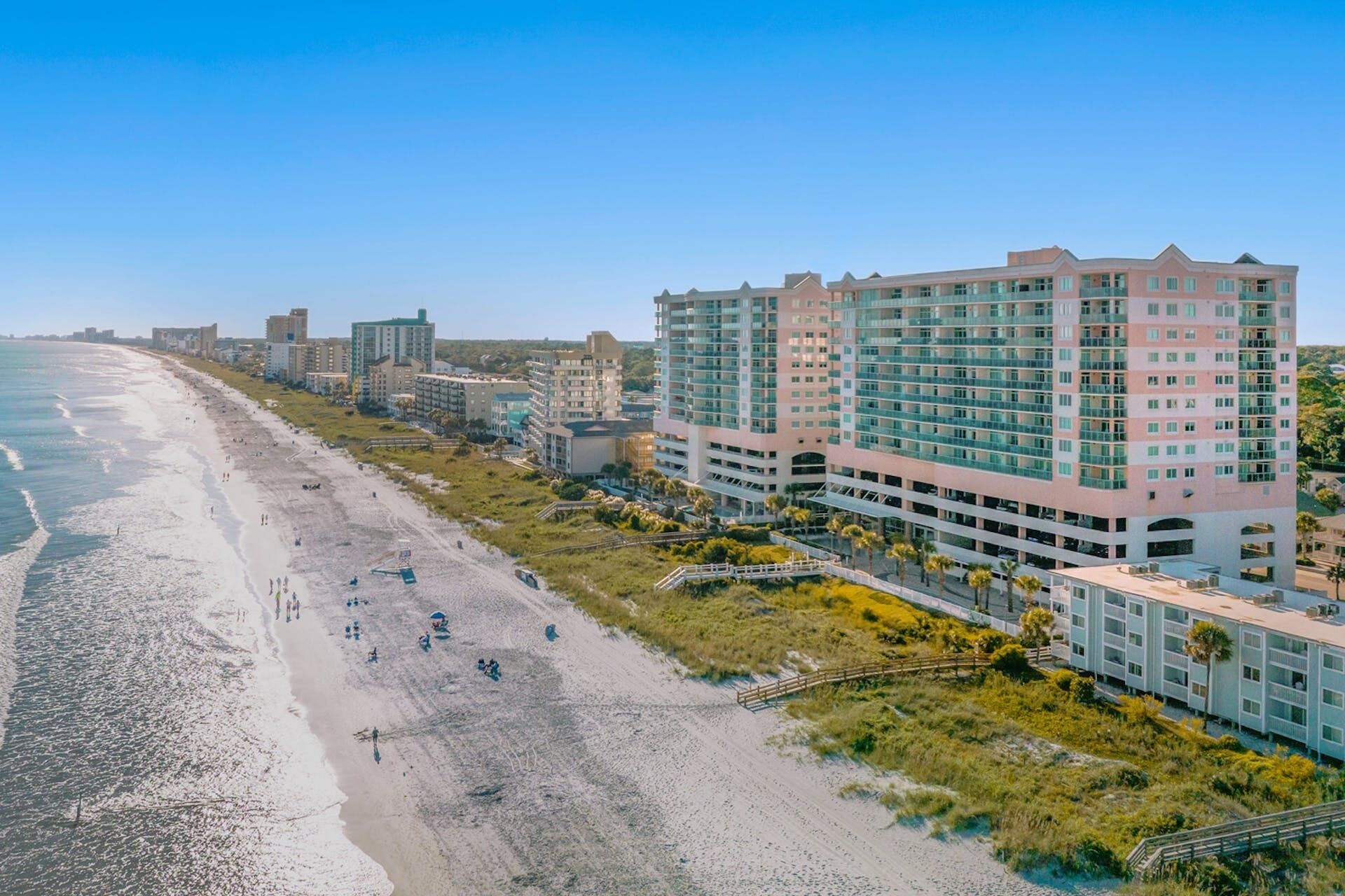 A beachfront hotel with multiple floors, a beach in the foreground, and a clear sky.