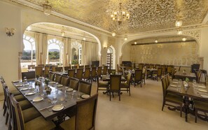 A spacious dining hall with ornate ceiling and chandeliers, large windows with curtains, and multiple dining tables set for guests. - The Jaibagh Palace (Amer)
