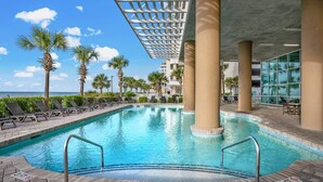 An indoor swimming pool with clear blue water, surrounded by lounge chairs and palm trees, under a covered area with a view of the ocean.