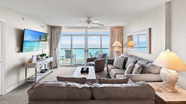 A spacious living room with a large sofa, a coffee table, and a TV mounted on the wall. There is a balcony with a view of the ocean. - Great oceanfront view from this 3 bedroom condo in the heart of NMB! | Crescent Keyes - 1205 (North Myrtle Beach)
