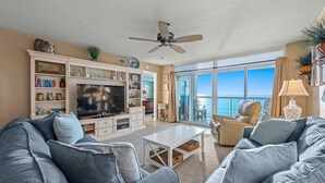A living room with a large flat-screen TV, a ceiling fan, and a view of the ocean through large windows. - Beautiful 3 bedroom condo with a great oceanfront view! | Crescent Keyes -  710 (North Myrtle Beach)