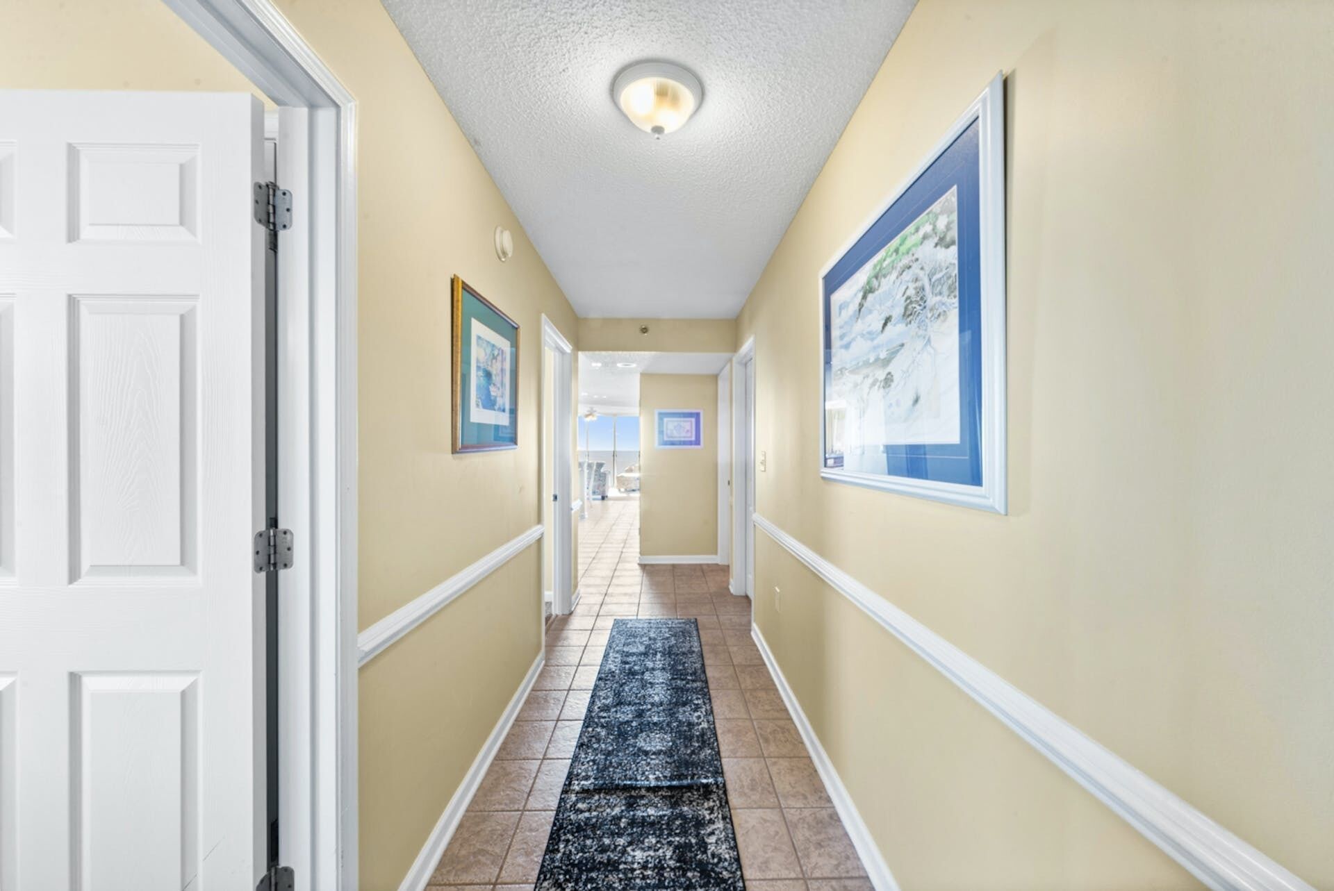 A hallway with beige walls, white trim, and a black and white rug. There are framed pictures on the walls and a white door on the left.