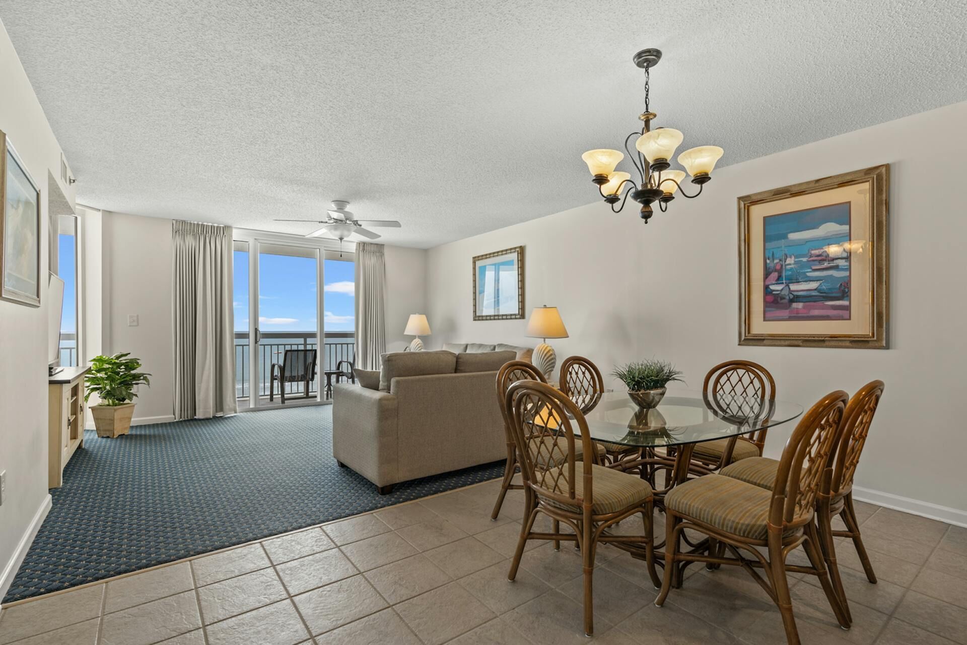 A hotel room with a balcony, a dining area with wicker chairs, a sofa, and a ceiling fan.