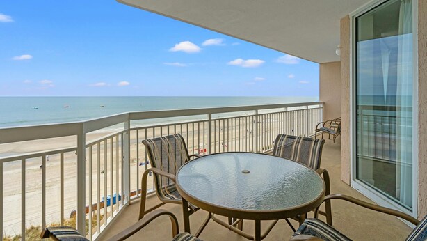A balcony with a glass table and chairs overlooking a beach.