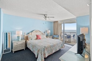 Une chambre d’hôtel comprenant un lit, deux tables de chevet avec des lampes, un ventilateur de plafond, une grande fenêtre donnant sur l’océan et un téléviseur posé sur un meuble.