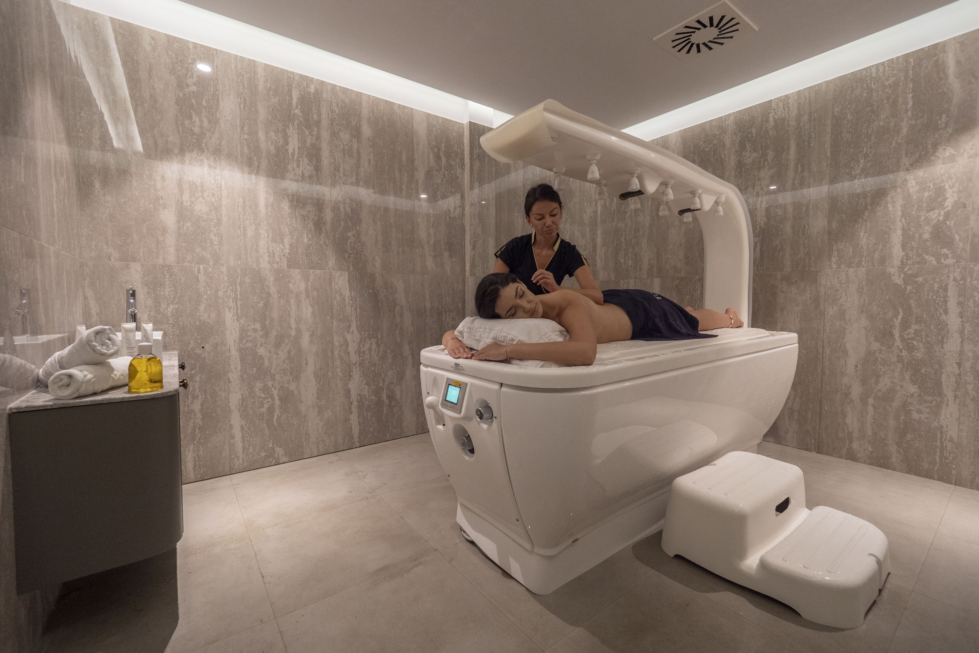 Un moderno cuarto de spa con una camilla de masaje, una mesita auxiliar con toallas y una botella, y un cabezal de ducha instalado en la pared.