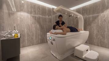 Un moderno cuarto de spa con una camilla de masaje, una mesita auxiliar con toallas y una botella, y un cabezal de ducha instalado en la pared.