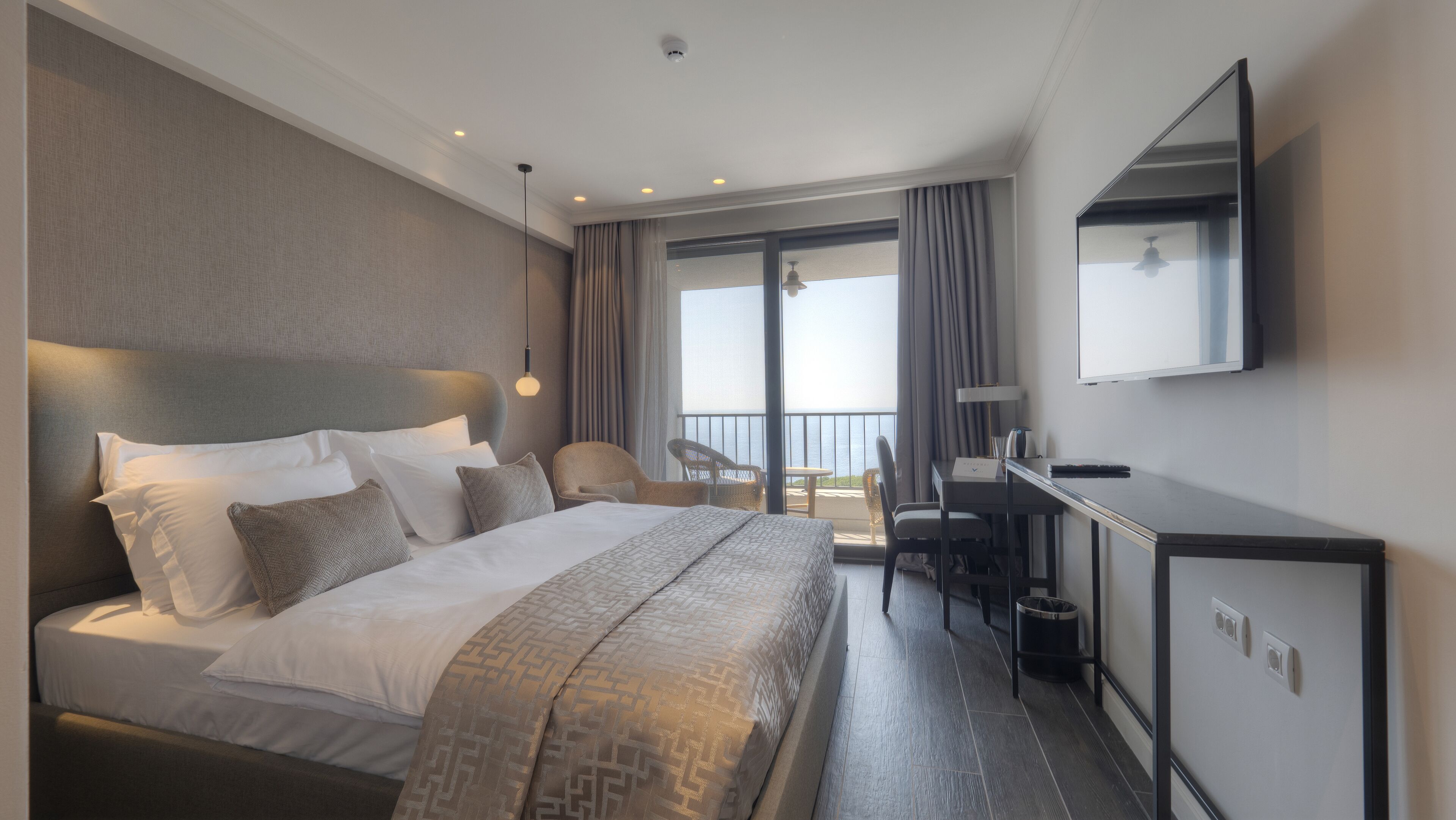 A hotel room with a large bed, a desk, a chair, a TV, and a balcony with a view of the sea.