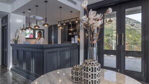 A reception area with a dark wood reception desk, multiple pendant lights, a vase with flowers, and a view of a mountainous landscape through large windows. - Vivid Blue Serenity Resort (Petrovac)