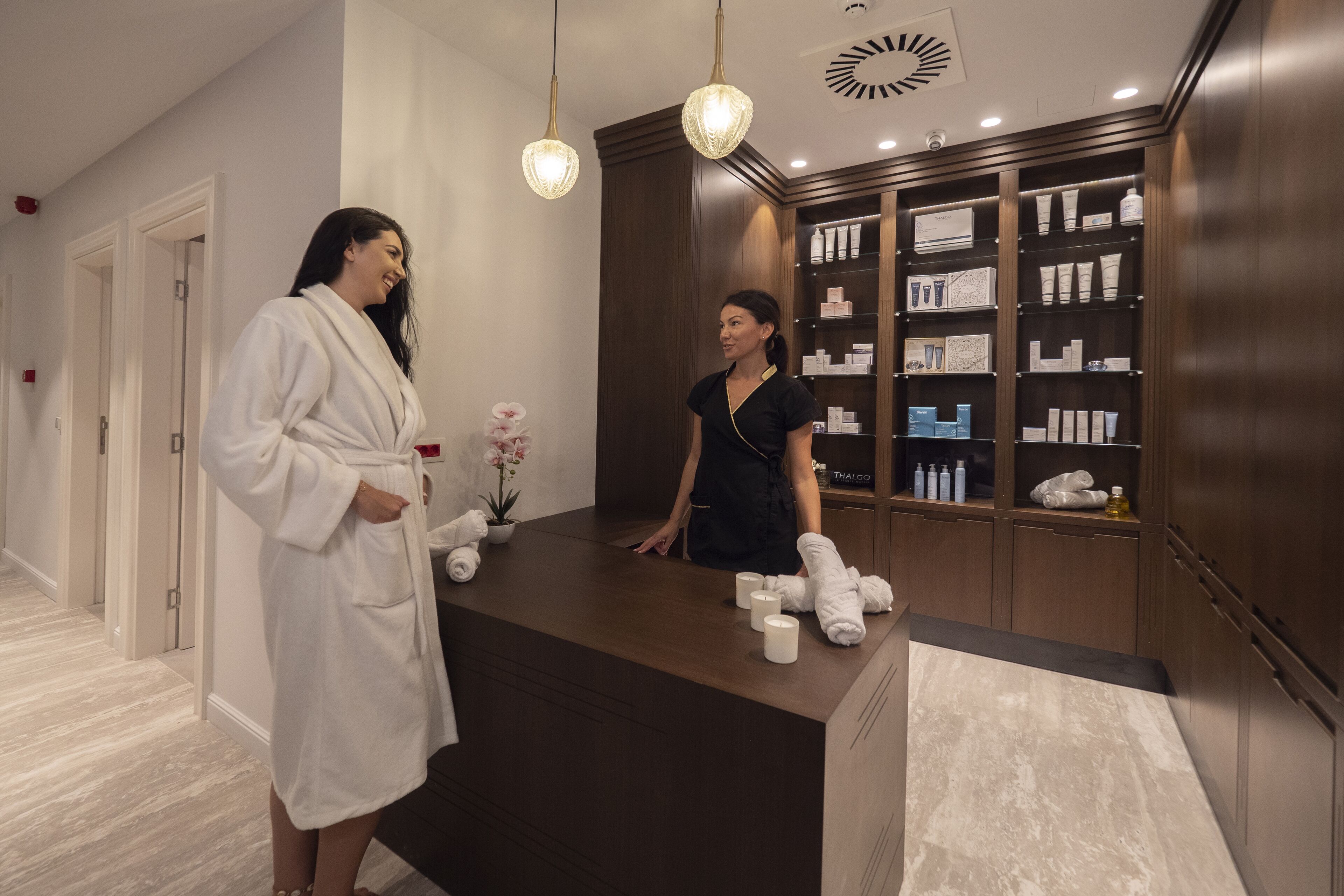 A reception area with a wooden reception desk, a white robe hanging on the wall, and a shelf with various toiletries.