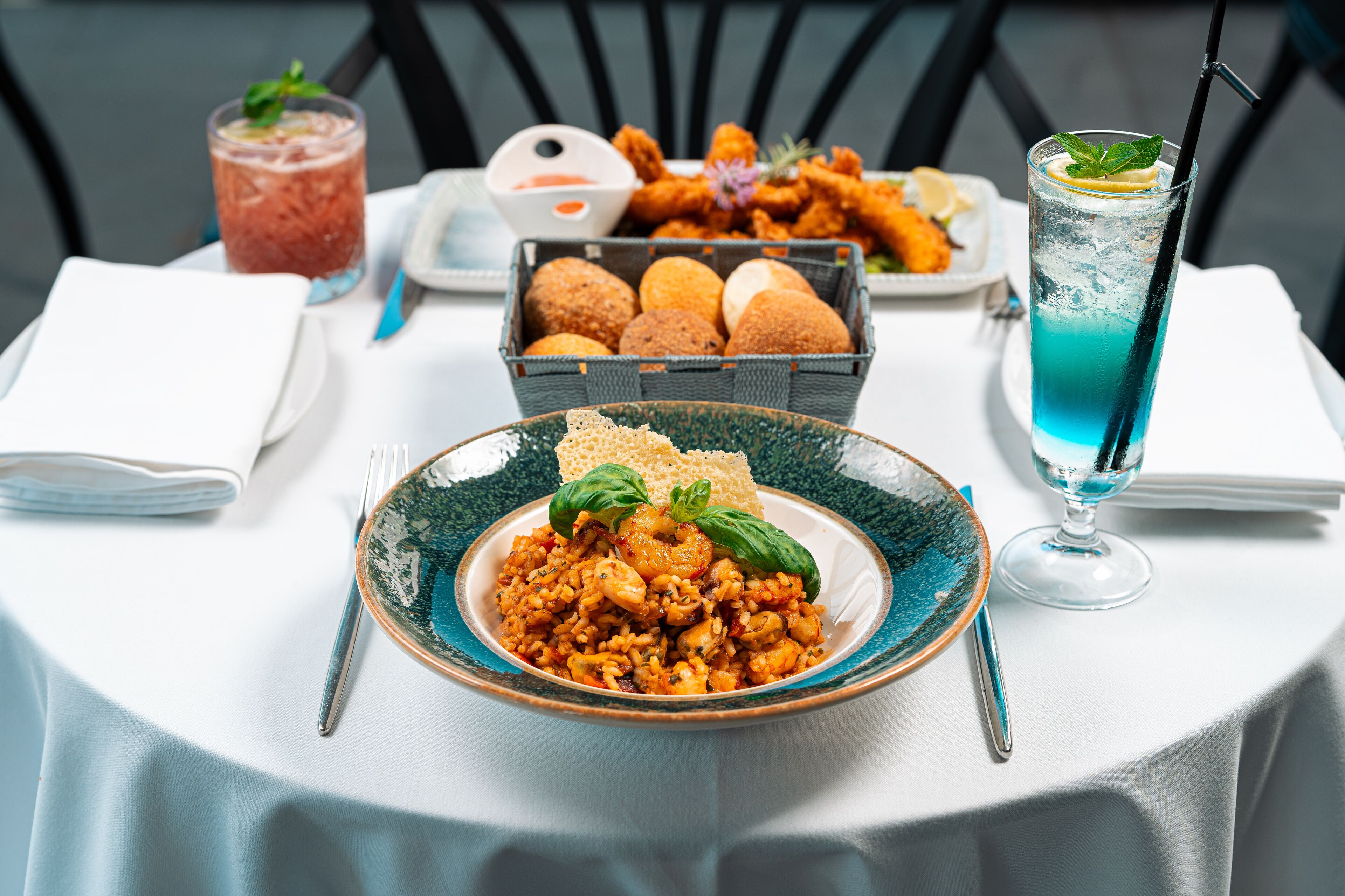 Una mesa dispuesta con un plato de mariscos, pan, una bebida y una cesta de panecillos.