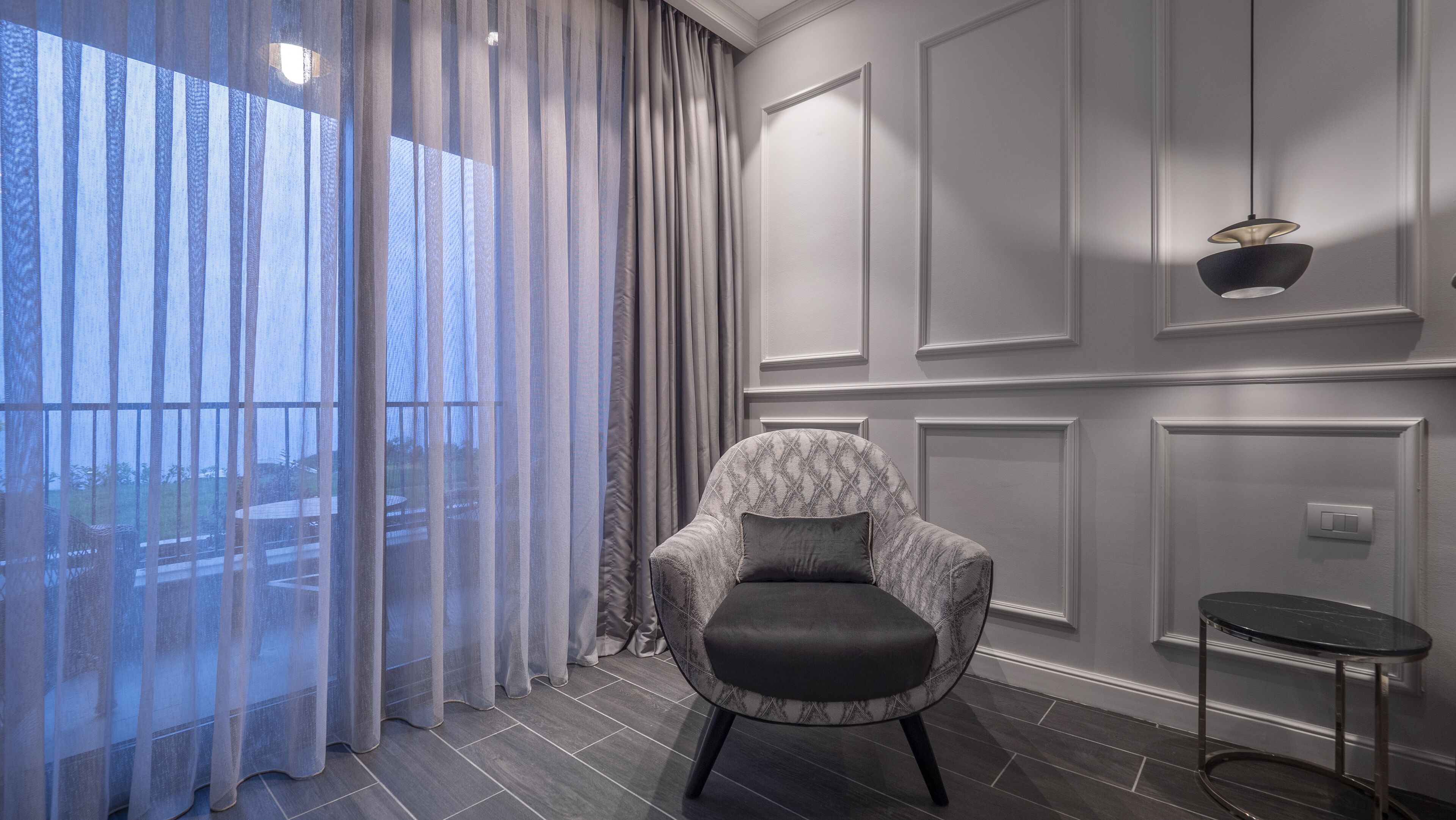 Une chambre moderne avec vue à travers de fines rideaux, un fauteuil à motifs, une petite table ronde et un luminaire fixé au mur.