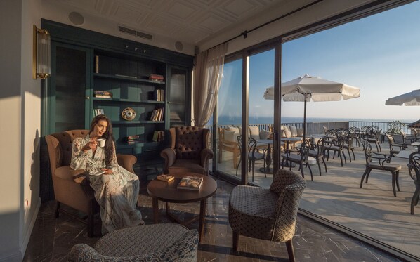 Una habitación con un gran ventanal, una estantería, un rincón de descanso con una mesa y un balcón con mobiliario exterior.