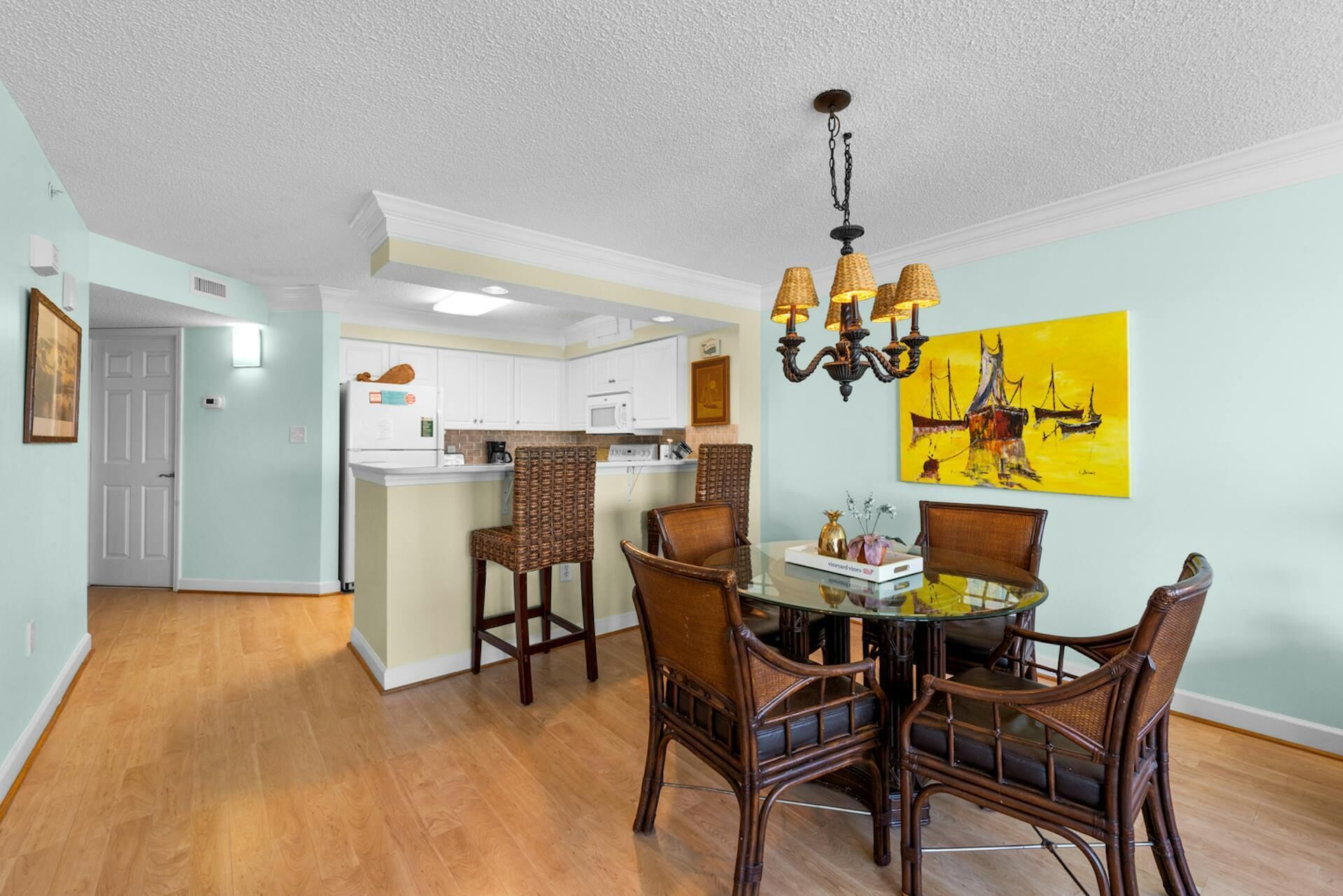 A dining area with a glass table surrounded by wicker chairs, a kitchen in the background, and a yellow painting on the wall.
