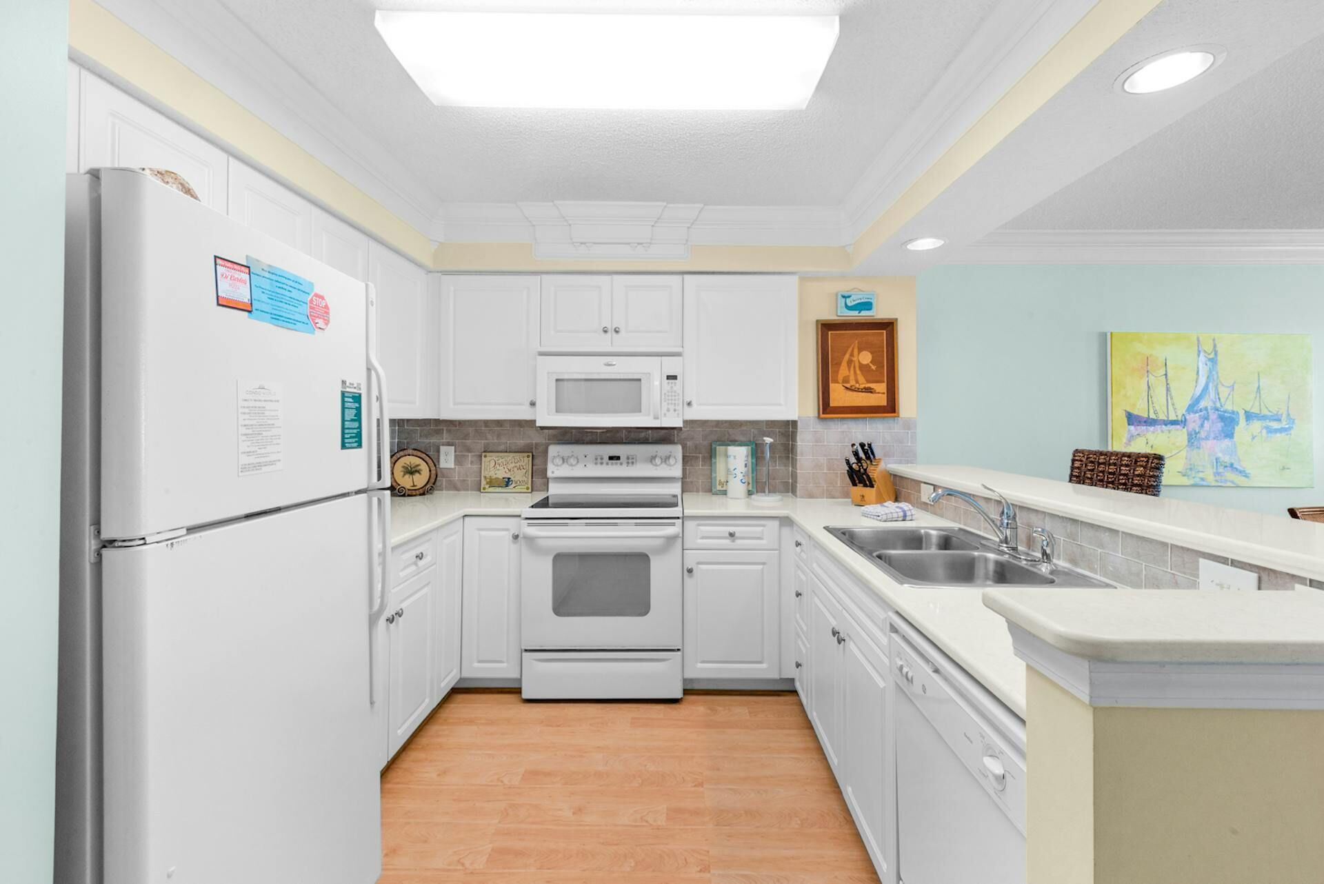 A modern kitchen with white cabinetry, a built-in oven, microwave, and dishwasher. There is a double sink and a wooden floor.