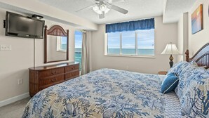Une chambre à coucher comprenant un lit, une commode, un ventilateur de plafond, un téléviseur, une fenêtre donnant sur l’océan et une lampe.
