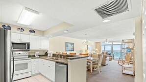 Een moderne keuken met witte kastjes, een ingebouwde magnetron en een vaatwasser. Er is een eethoek met een tafel en stoelen, en een uitzicht op de oceaan door grote ramen.