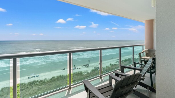 Een balkon met uitzicht op zee en strand, met twee Adirondack stoelen en een glazen hekwerk.