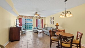 A living room with a sofa, a dining table with chairs, a ceiling fan, and a view of a building through a window.