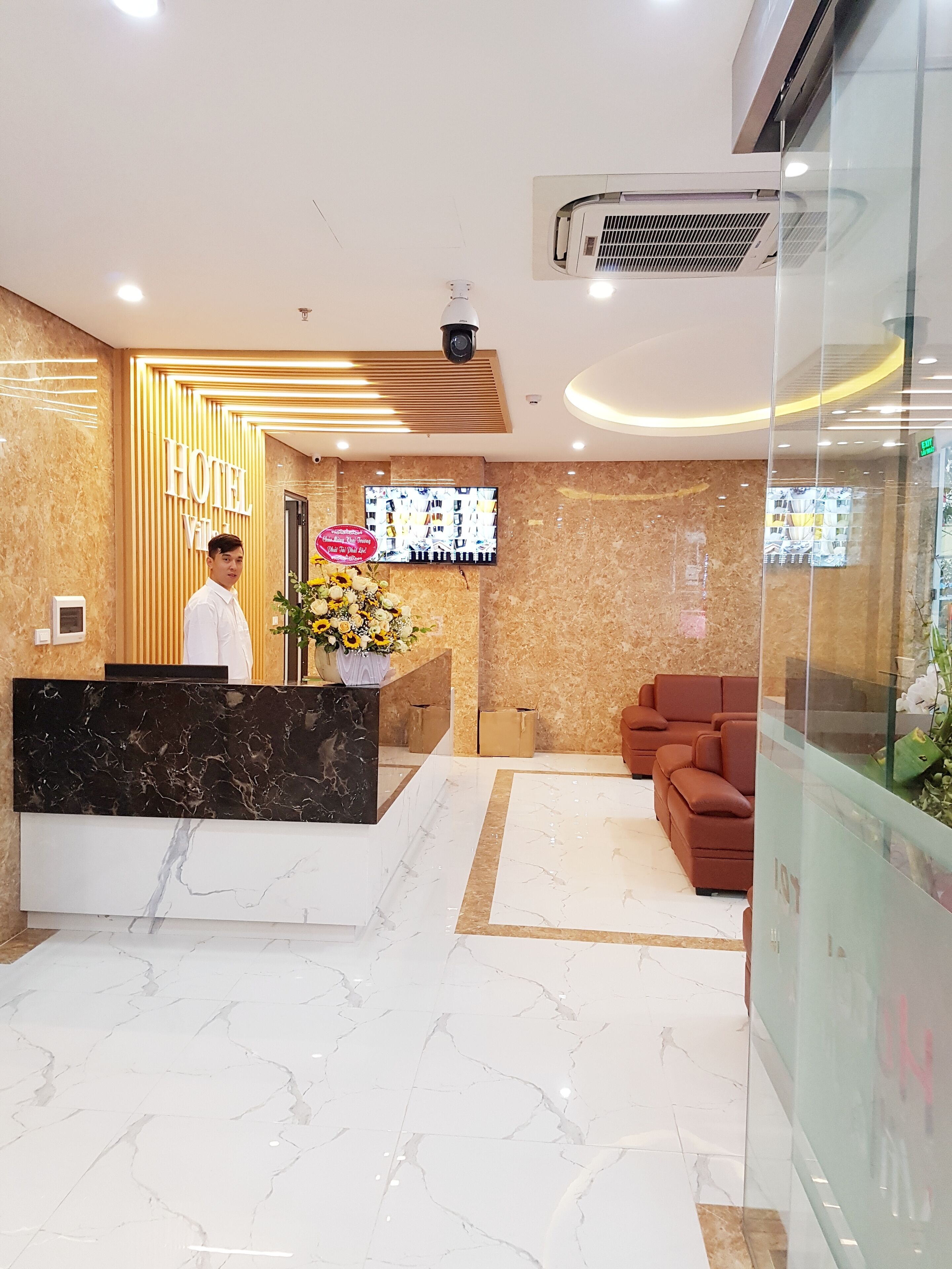 Zona de recepción de hotel con un mostrador de mármol, un sofá y una pantalla de televisión mostrando imágenes.