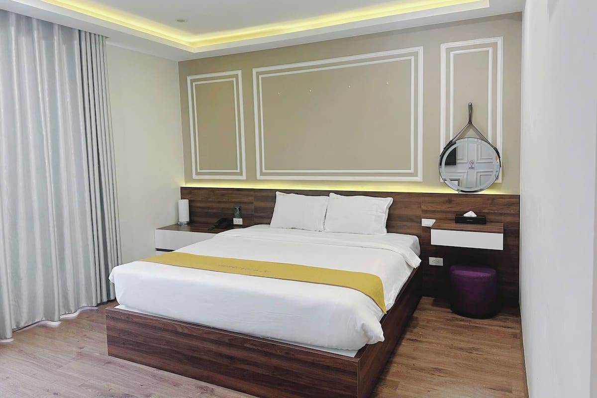 A modern hotel room with a wooden bed, a bedside table with a lamp, a mirror, and a small purple stool.