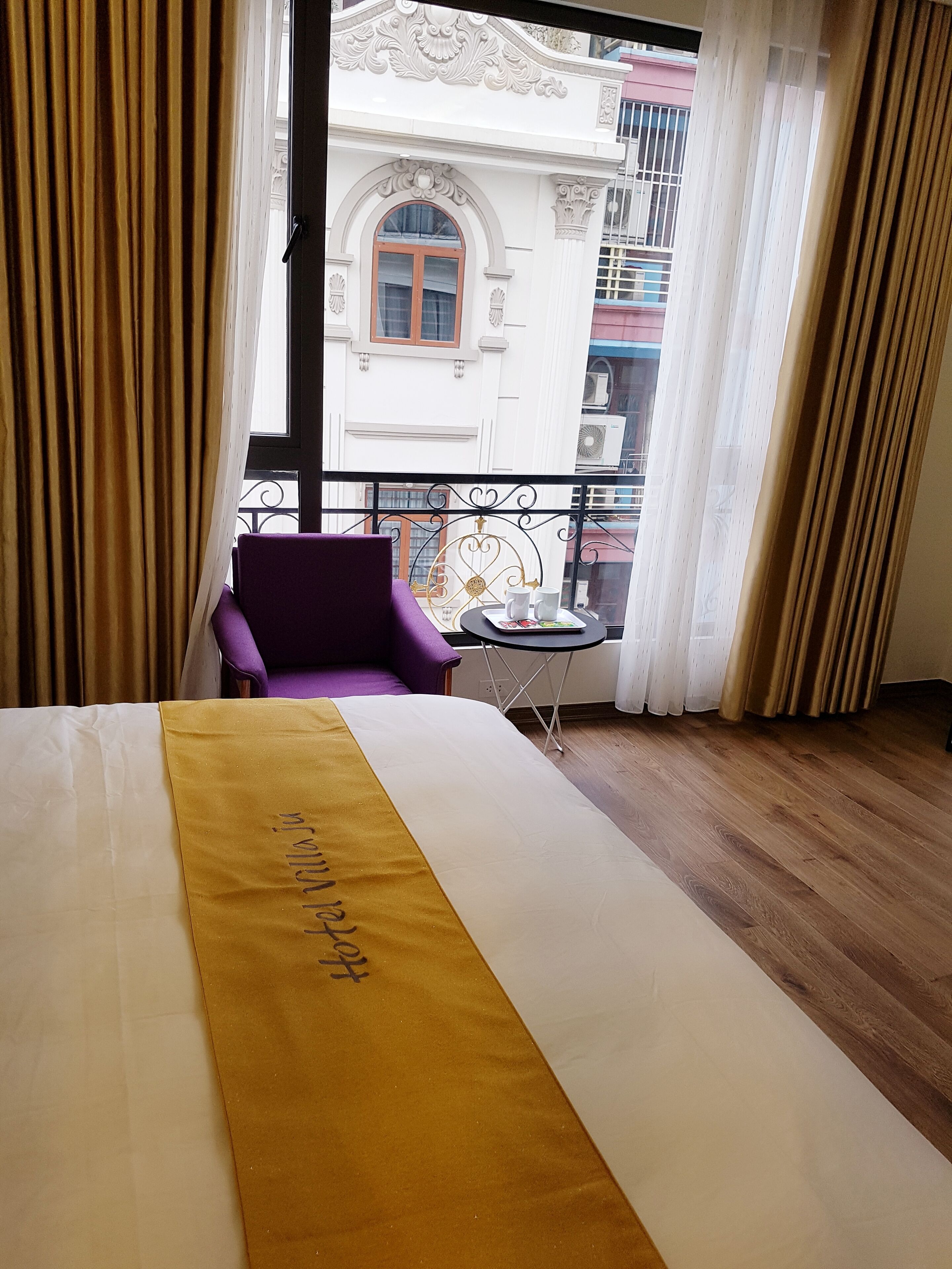 Une chambre d’hôtel avec un lit, un fauteuil violet, une petite table avec un plateau, et une vue sur le bâtiment situé à l’extérieur.