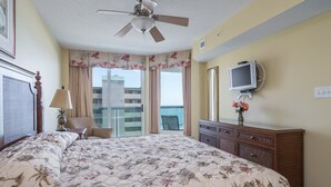 Een hotelkamer met een groot bed, een plafondventilator, een televisie, een dressoir en uitzicht op de oceaan.