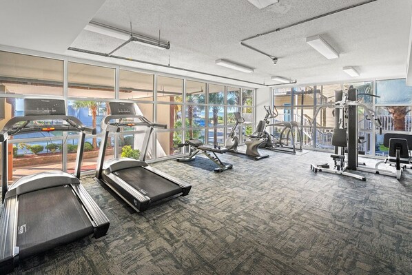 A modern gym with treadmills, elliptical machines, and weight equipment. Large windows offer views of a landscaped area and a body of water. - 3-bedroom, 3-bathroom, 6th floor condo in Crescent Beach | Malibu Pointe - 604 (North Myrtle Beach)