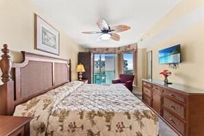 Een hotelkamer met een bed, een plafondventilator, een televisie, een dressoir en een raam met uitzicht op gebouwen.