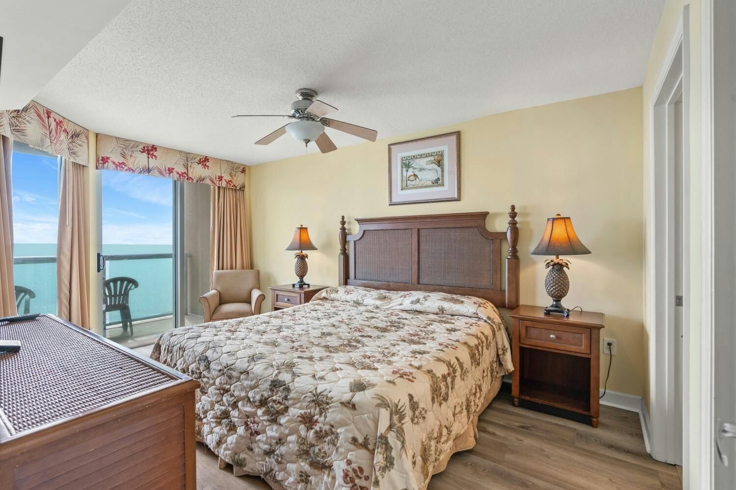 Une chambre d’hôtel avec un lit, deux tables de chevet, une chaise, un ventilateur de plafond et une vue sur l’océan.