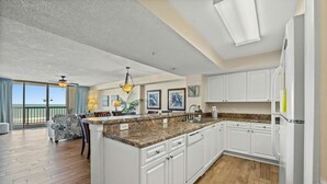 A spacious kitchen with white cabinets, a granite countertop, and a large island. There is a dining area with a sofa and a table, and a balcony with a view of the ocean. - Oceanfront 3-bedroom, 3-bathroom condo, jetted tub, private balcony | Ocean Bay Club - 308 (North Myrtle Beach)