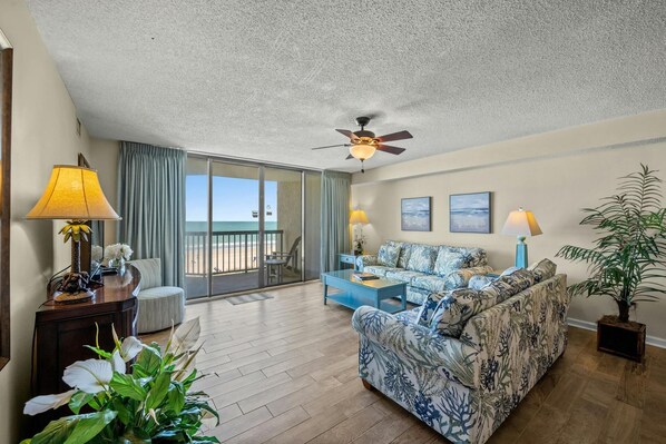 A spacious living room with a large sofa, a coffee table, and a view of the beach through a sliding glass door.