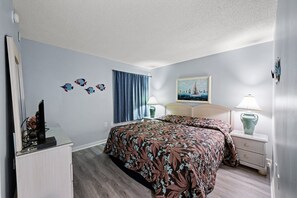 A hotel room with a bed, a desk, a lamp, and a painting on the wall. - 4th floor charmer with ocean views, 1-bedroom, 1-bath, Ocean Drive condo | Ocean Bay Club - 406B (North Myrtle Beach)
