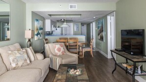 A living room with a sofa, coffee table, and TV stand. There is a dining area with chairs and a table. The room has a kitchen in the background.