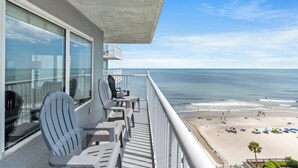 Een balkon met twee Adirondack stoelen en een tafel, met uitzicht op een strand met parasols en mensen.