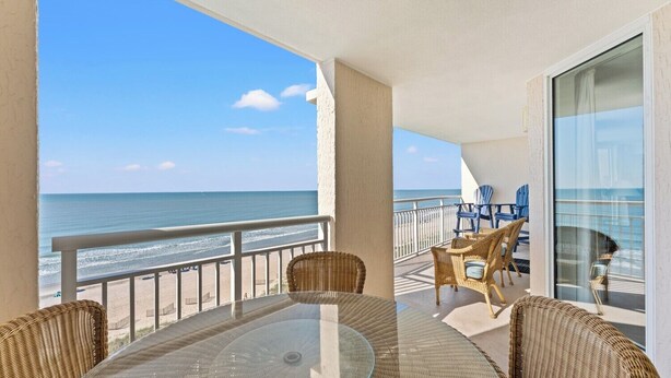 A balcony with a glass table and wicker chairs, overlooking a beach and ocean. - Tastefully decorated three-bed, three-bath oceanfront condo | South Shore Villas - 702 (North Myrtle Beach)