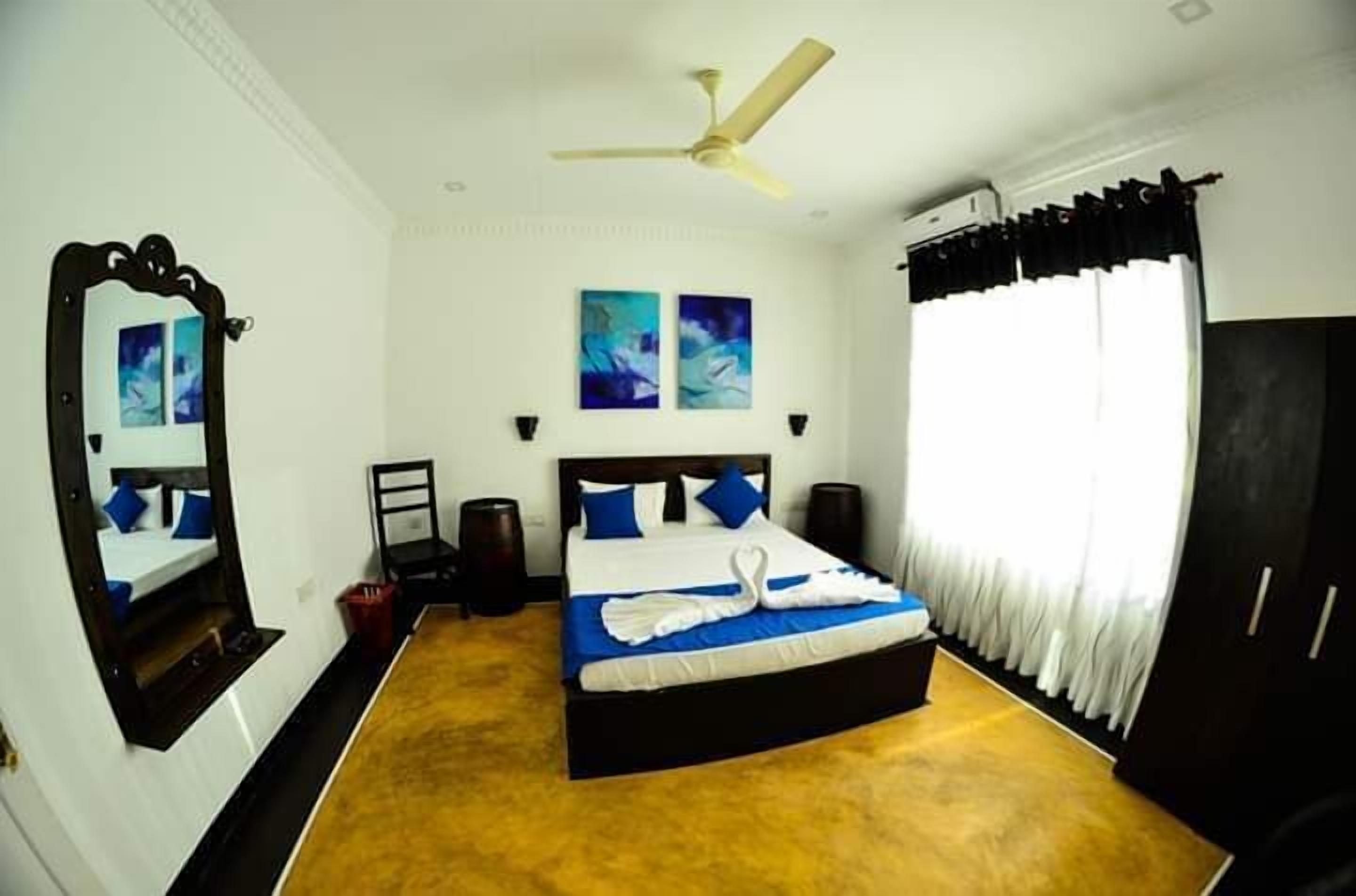 A hotel room with a large bed, a ceiling fan, two wall-mounted lamps, a mirror, and two paintings on the wall.